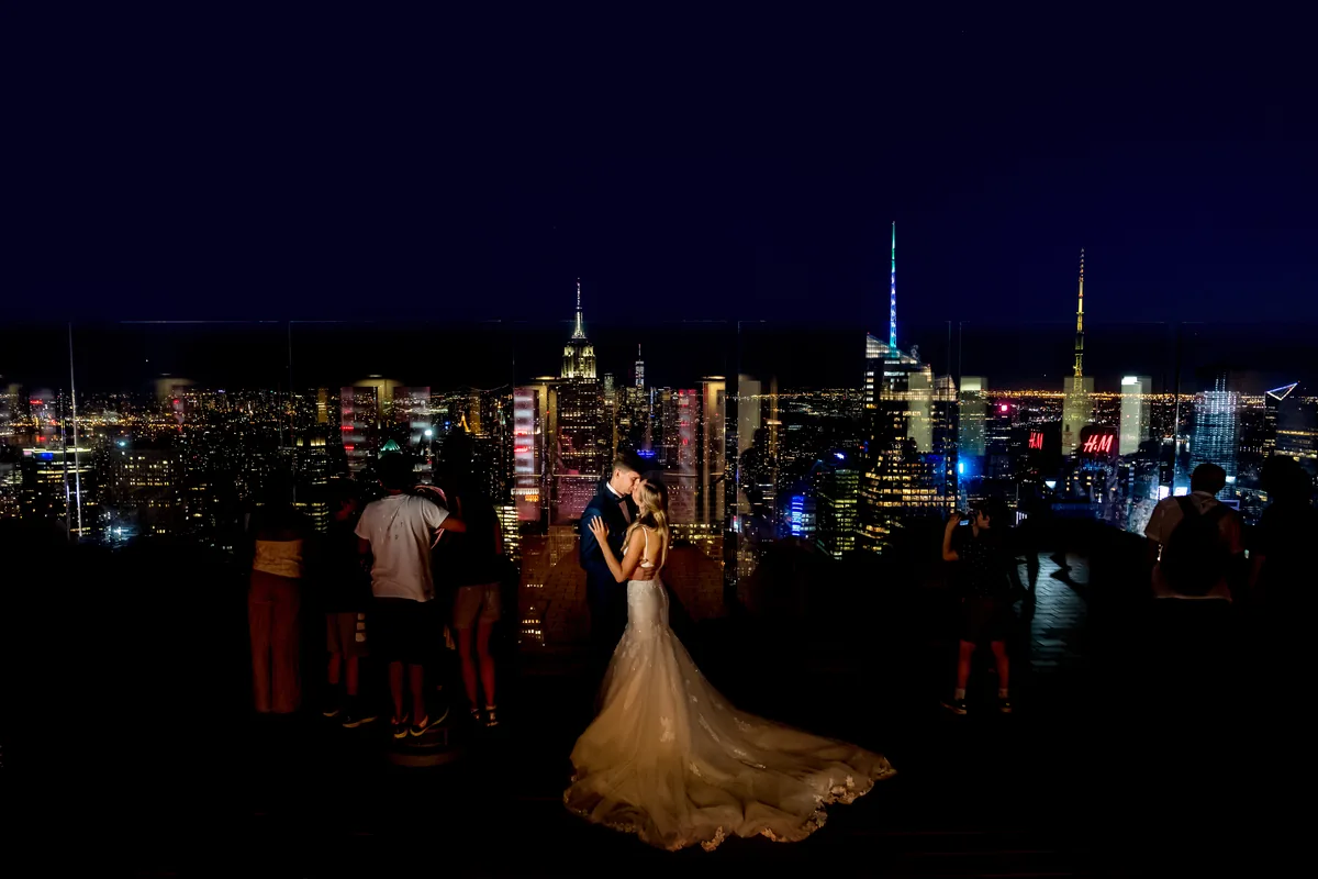 Miri sărutându-se la balustrada de sticlă, siluete de turiști și Manhattanul luminat