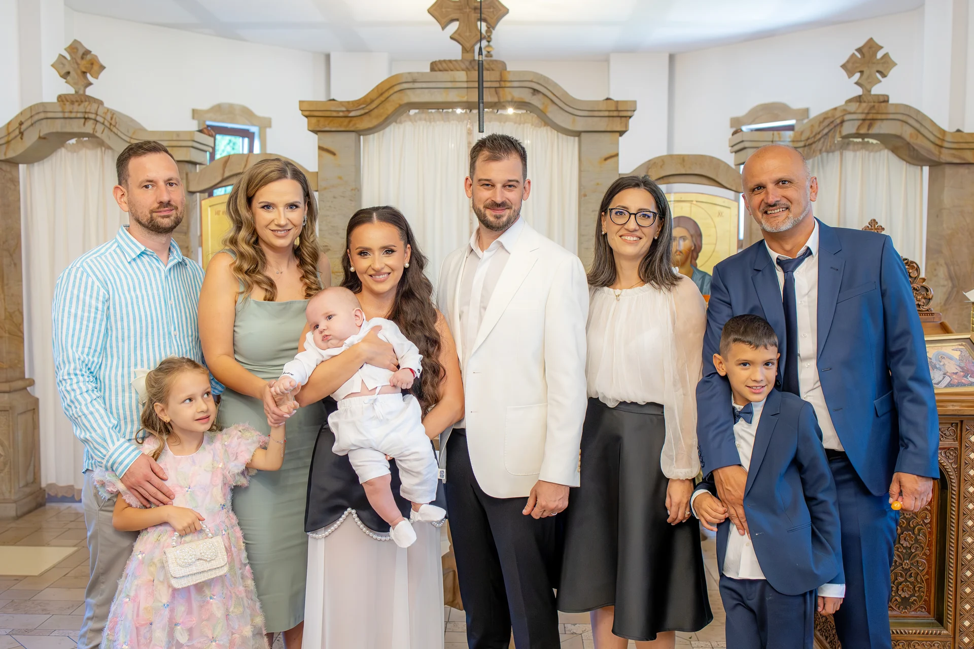 Familie de opt, cu bebelușul în brațe, în fața iconostasului de piatră cu cruce