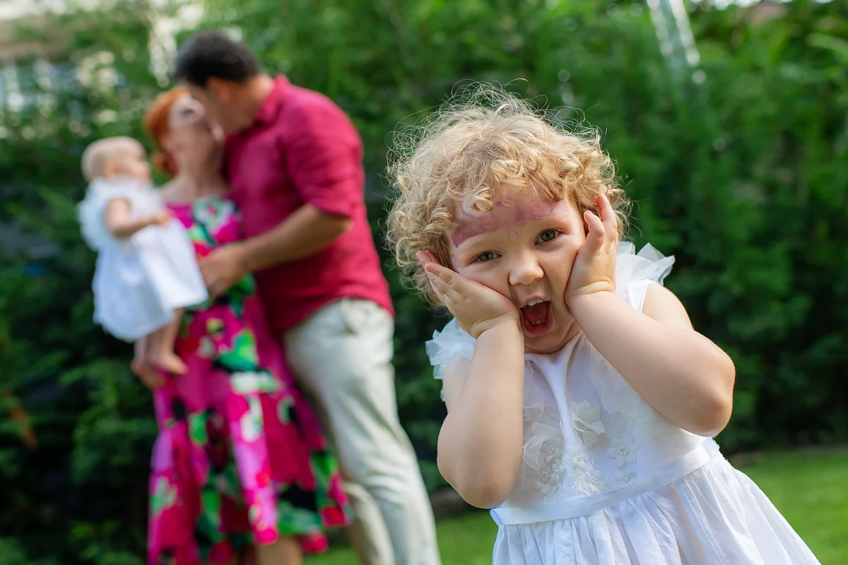 Fetița surprinsă, cu mâinile pe obraji, în timp ce părinții se sărută ținând bebelușul în grădină — moment candid la un botez lângă Cluj-Napoca
