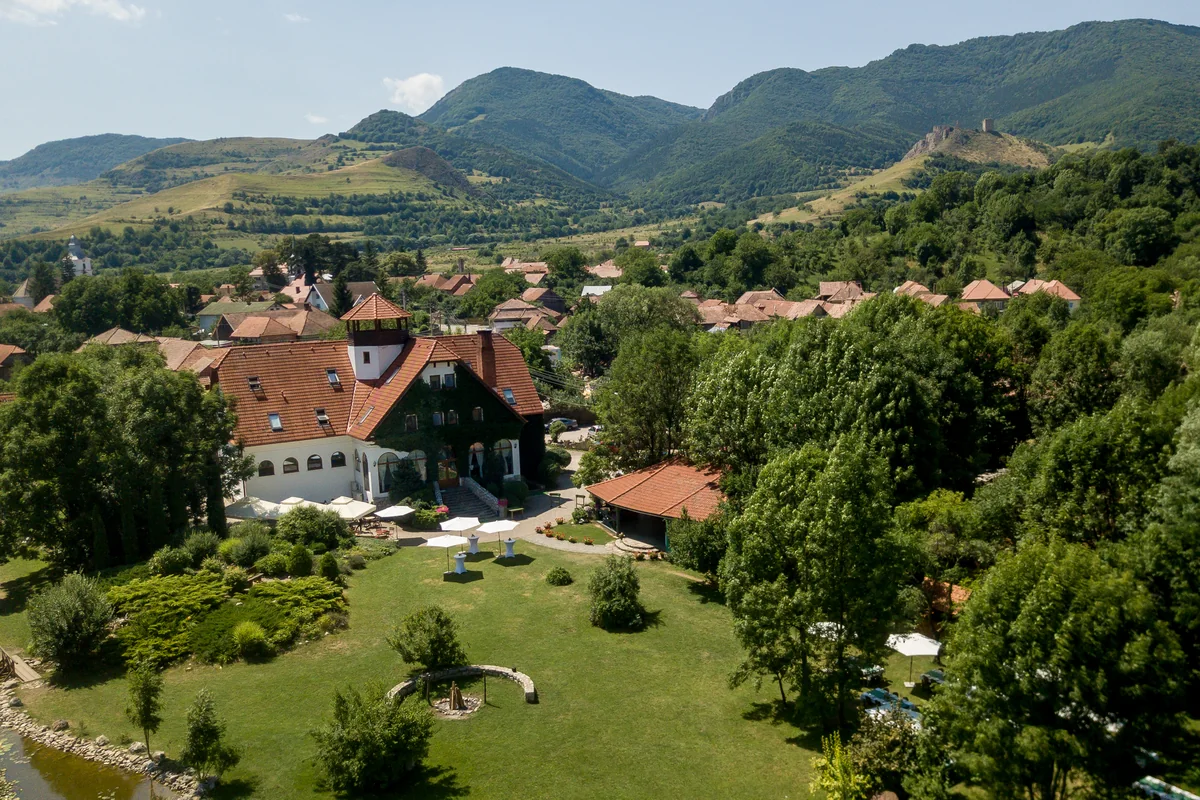 Vedere din dronă a Conacului Secuiesc — clădirea cu turn, pajiștea verde și satul, cu masivul Colțești și ruinele cetății în fundal