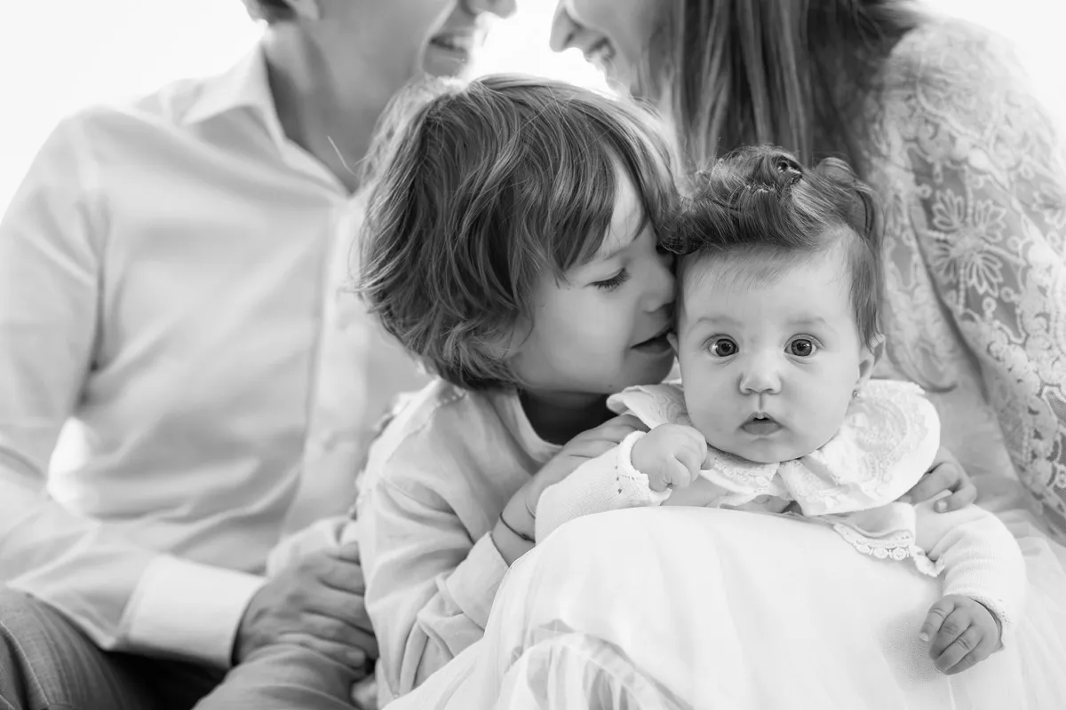 Surioară sărutând-o pe frunte pe bebelușa de la botez, în brațele familiei, portret alb-negru