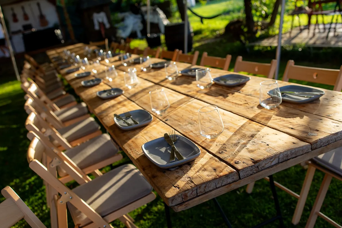 Masă lungă rustică din lemn, aranjată în aer liber cu farfurii, tacâmuri și pahare pentru invitații de la botez — costul care se acoperă din daruri