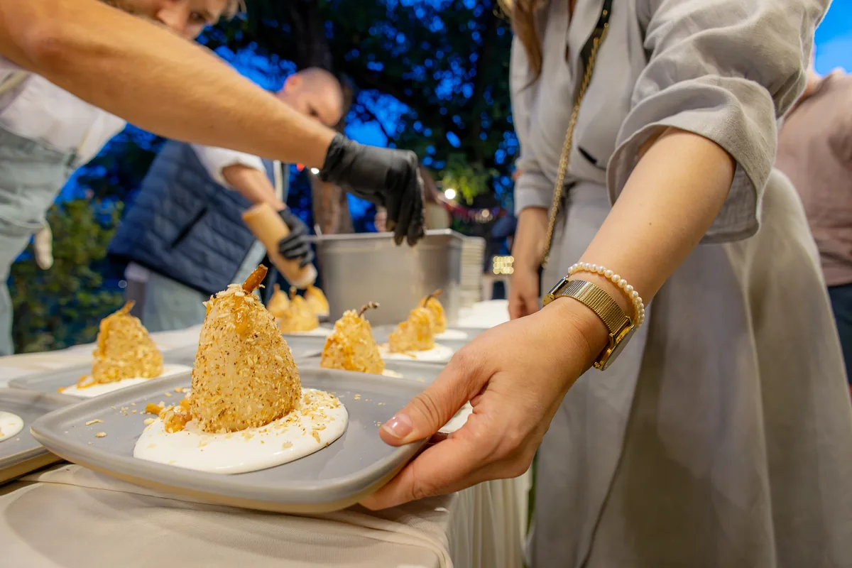 Catering la botez — desert din pară servit pe farfurie unei invitate, seara în grădină; meniul e reperul de cost al petrecerii