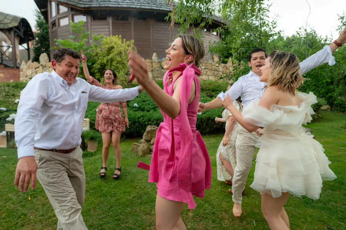 Invitați dansând desculți pe iarbă în ploaie, mireasa în rochie scurtă albă