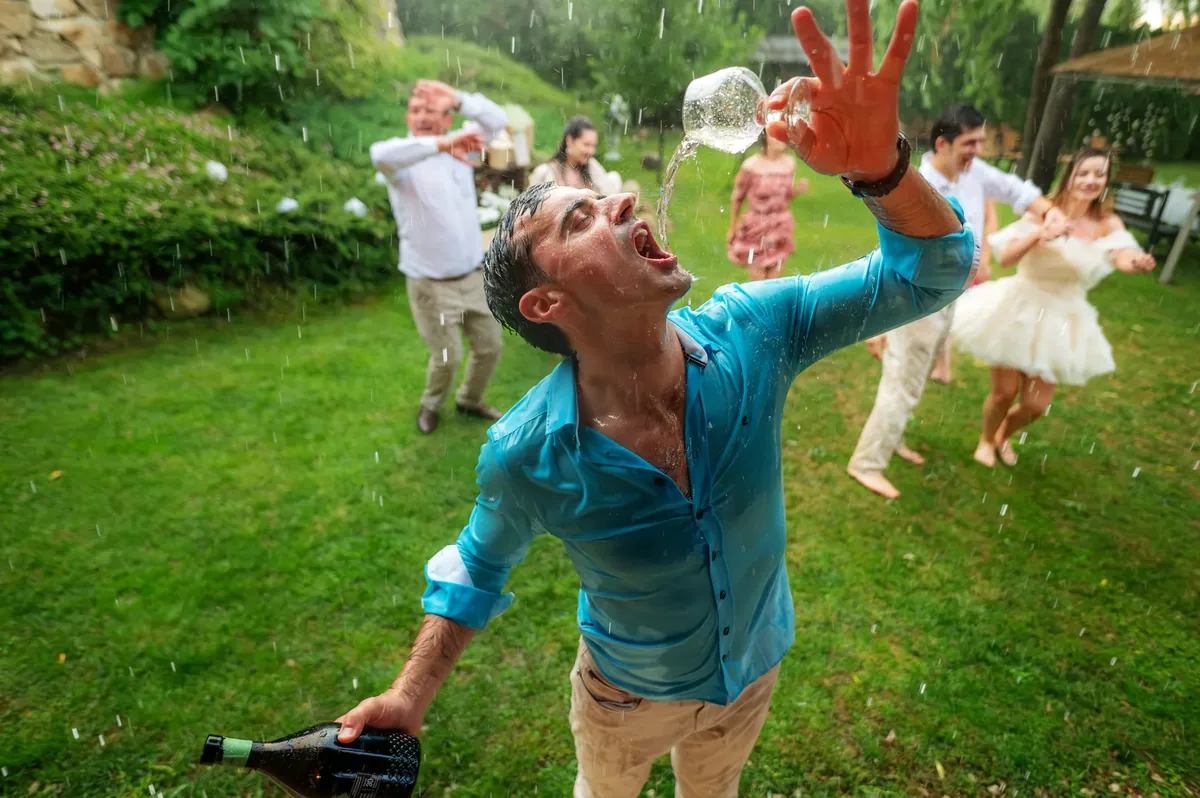 Un invitat în cămașă udă râde, vărsând șampanie din pahar, sub ploaia torențială