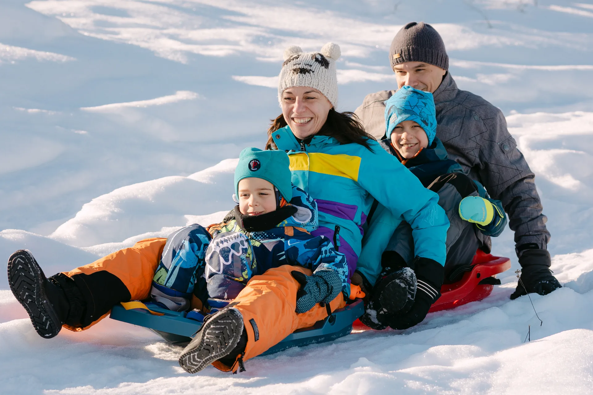 Familia îngrămădită împreună pe sanie în zăpadă, râzând, în costume colorate