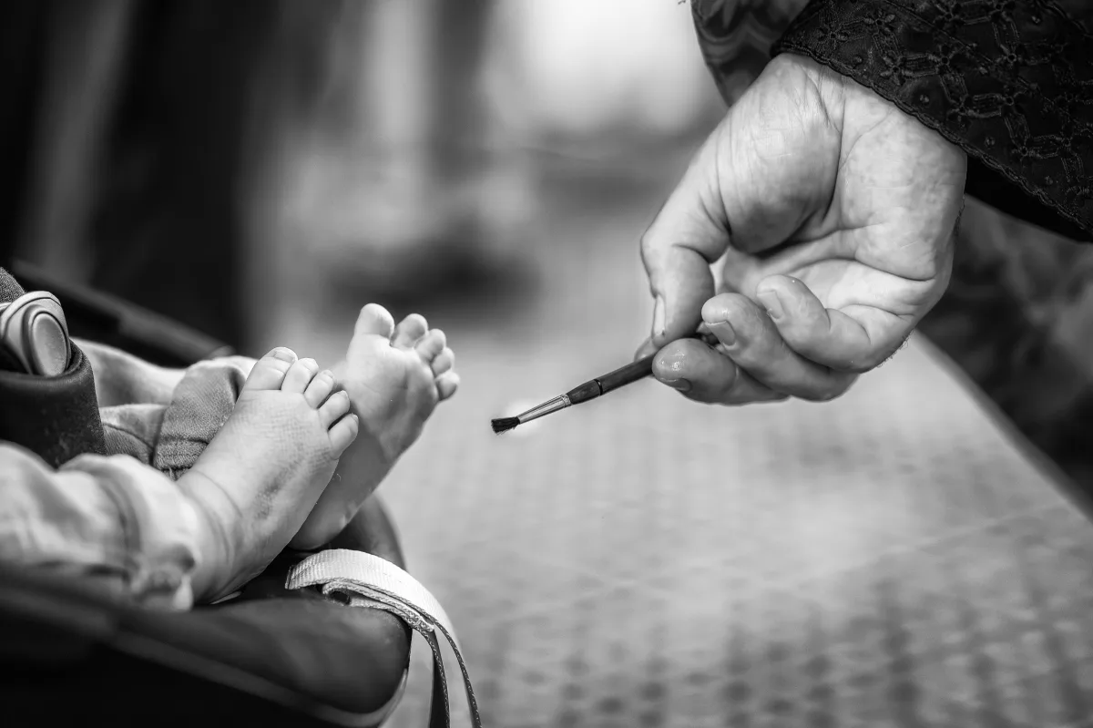 Preotul atinge cu pensula de mir tălpile bebelușului la botez — cadru alb-negru în biserică