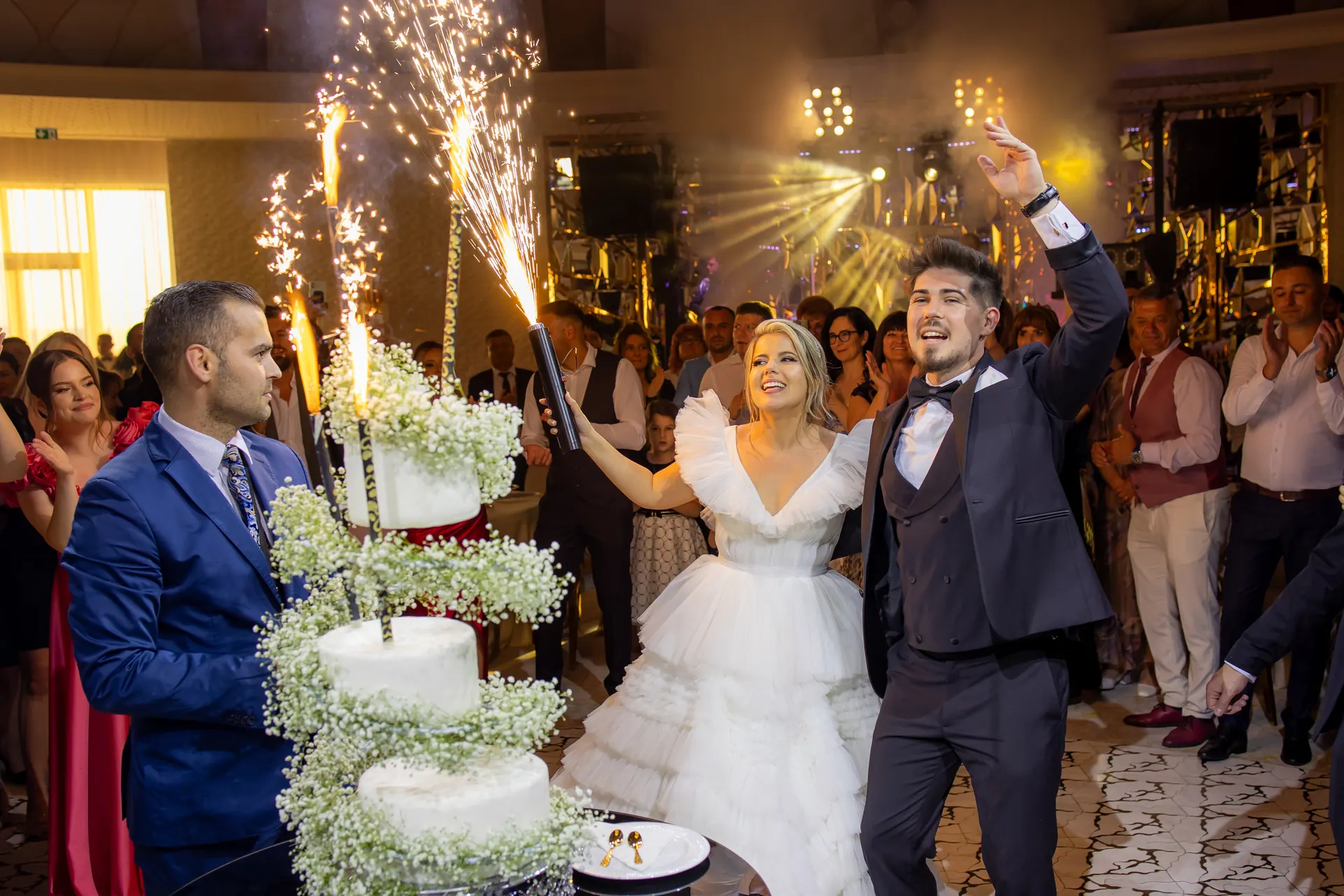 Mirii lângă tortul cu trei etaje și artificii, invitații aplaudând în jur