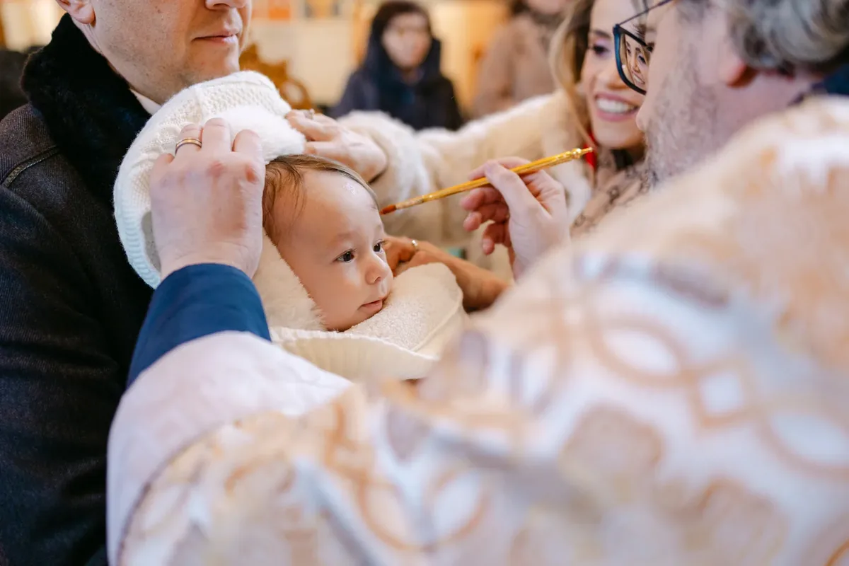 Preotul unge cu pensula fruntea bebelușului înfășurat în alb, iar mama zâmbește alături — botez la Biserica Sfântul Gheorghe din Grigorescu