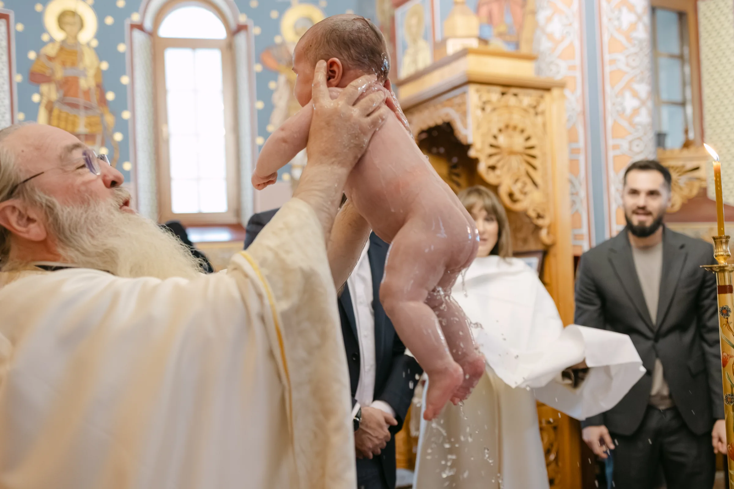 Preotul ridicându-l pe bebeluș deasupra cristelniței, cu apa șiroind, invitații privind