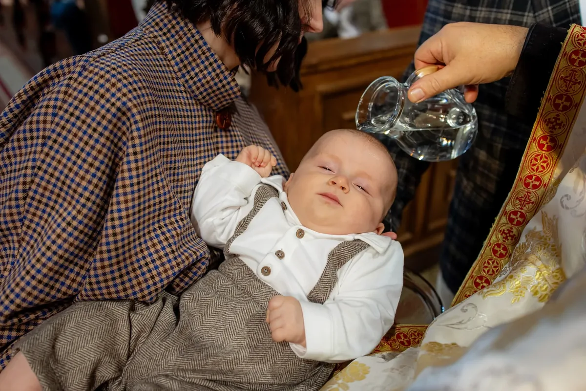 Apă turnată dintr-o carafă de sticlă pe creștetul bebelușului îmbrăcat, ținut în brațe lângă veșmântul preotului